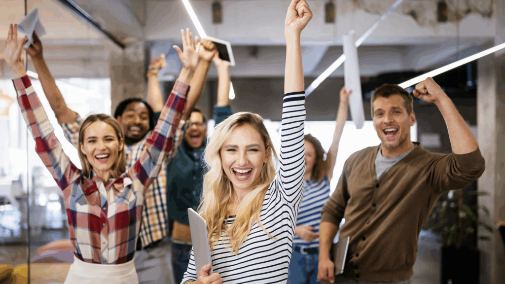 Group of diverse people smiling, raising their arms, and cheering together to celebrate a big achievement.
