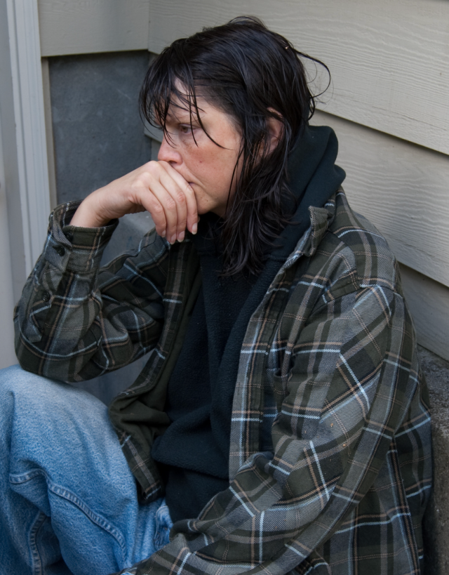 A woman struggling with drug addiction sits on the floor in front of a closed door, her head in her hands, looking exhausted and overwhelmed.
